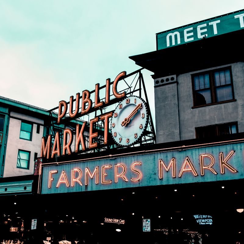 A public market with neon signs. The main sign reads "Public Market Farmers Market," and there's a clock above the "Public Market" sign.