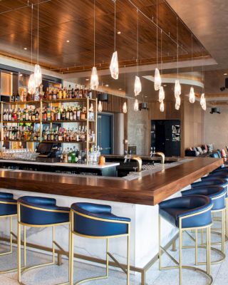 A modern bar with a wooden countertop, blue chairs, a well-stocked liquor shelf, and elegant pendant lights with a spacious seating area.
