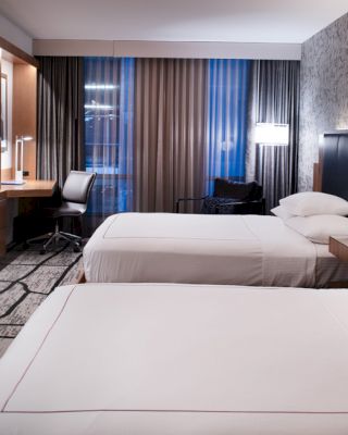 The image shows a modern hotel room with two beds, a TV mounted on the wall, a desk and chair, and nightstands with lamps.