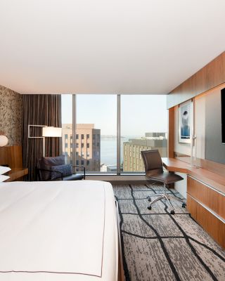 A modern hotel room with a large bed, flat-screen TV, desk, chair, and a window offering a cityscape view.