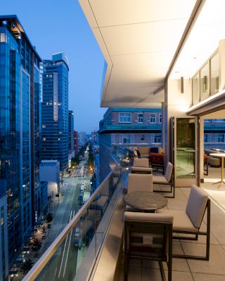 A modern balcony overlooks an urban cityscape with tall buildings. The balcony has seating and tables, and large windows provide indoor access.