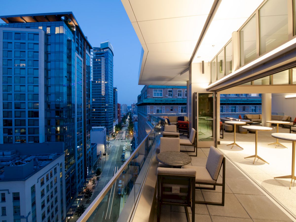 The image depicts a modern cityscape viewed from a building balcony or terrace at dusk, featuring tall glass buildings, tables, and seating.