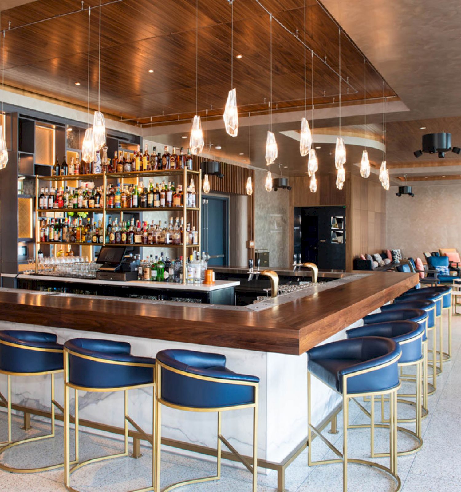 A modern bar with blue and gold stools, overhead lighting, and a fully stocked back wall. Adjacent seating areas are visible in the background.