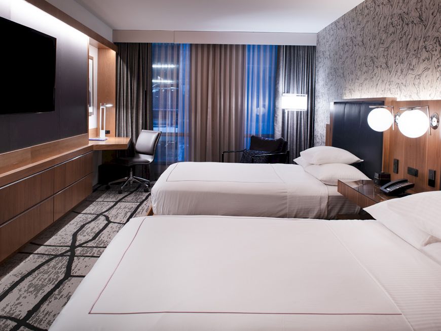 This image shows a modern hotel room with two double beds, a large TV, a desk with a chair, and curtains covering a window.