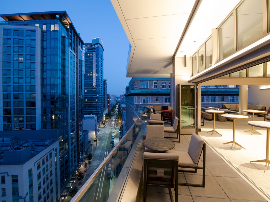 A modern, upscale balcony overlooks a cityscape at dusk, with tall buildings, a street below, and seating areas for dining or relaxation.
