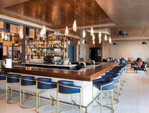 A modern bar with hanging lights, blue bar stools, and a fully stocked shelf of alcohol bottles behind the counter, in a spacious lounge area.