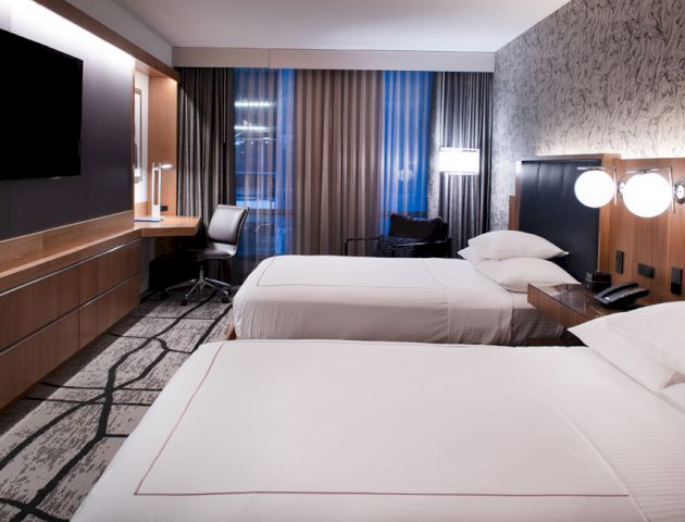 A modern hotel room features two beds, a flat-screen TV, a work desk, and stylish decor with patterned wallpaper and contemporary lighting.