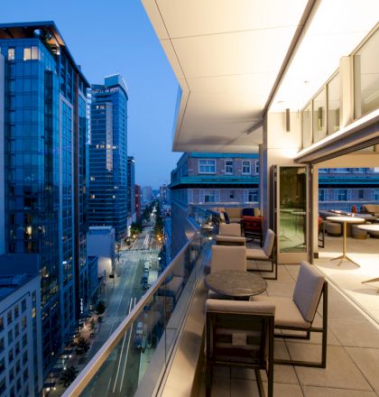 A modern balcony overlooking a cityscape at dusk, with seating and tables, tall buildings, and a street below with lit windows and streetlights.