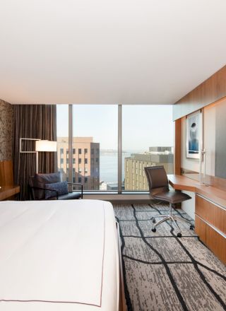 A modern hotel room features a neatly made bed, a large TV, a desk with a chair, a cozy seating area, and a window displaying an urban view.