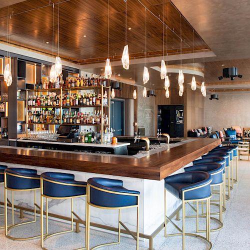 Modern bar area with a wooden counter, blue bar stools, hanging lights, and a large shelf of liquor bottles behind the bar.