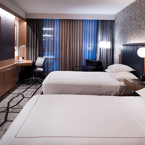 The image shows a modern hotel room with two beds, a large TV, a desk with a chair, and contemporary decor.