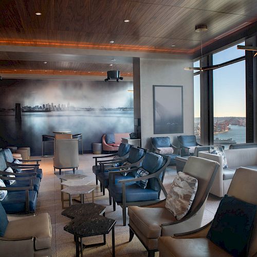 A modern lounge with stylish seating overlooks a picturesque water view. The room features a large mural of a city skyline and ample natural light.