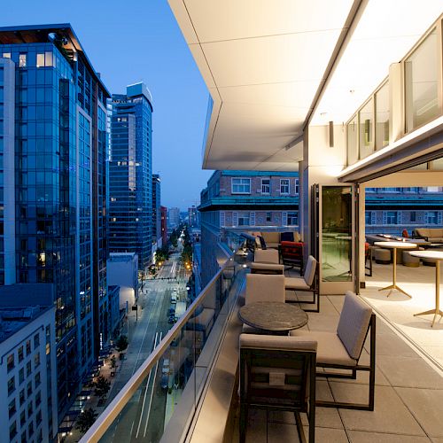 A modern cityscape view from a rooftop terrace with seating and tables, overlooking tall buildings and a street below during twilight.