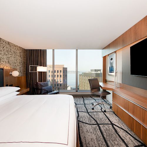 A modern hotel room with a large bed, a TV, a desk, a picture window with a view of buildings and water, and contemporary decor.