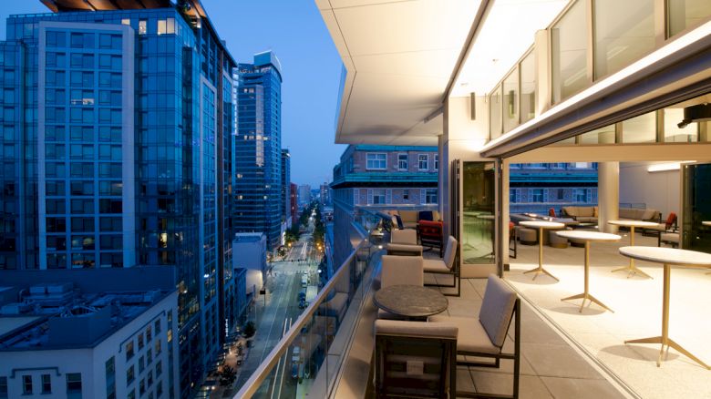 A modern outdoor balcony at dusk with tables and chairs, overlooking a cityscape of tall buildings and a well-lit street below.