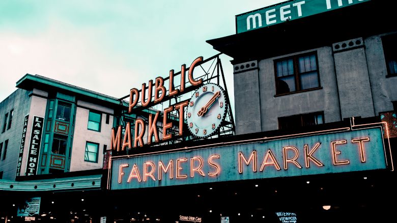 The image shows a building with a neon "Public Market" clock sign and "Farmers Market" below, indicating a vibrant marketplace setting.