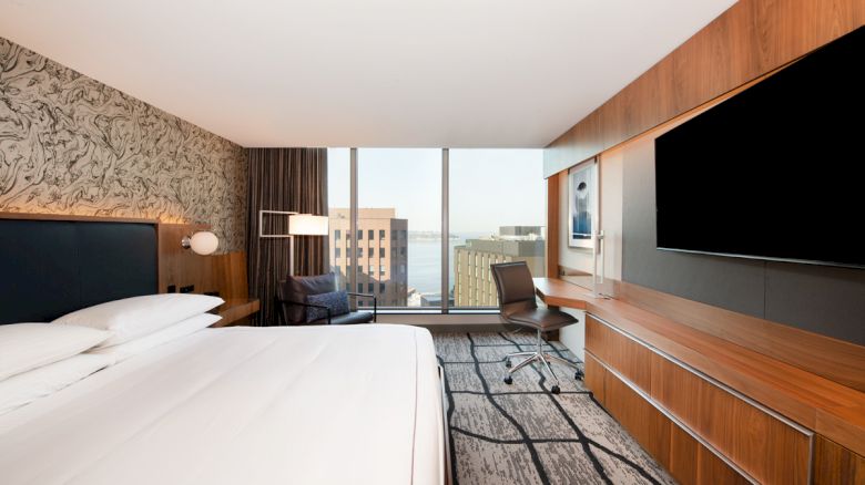 A modern hotel room features a large bed, desk, chair, flat-screen TV, and floor-to-ceiling window with a city view in the background.