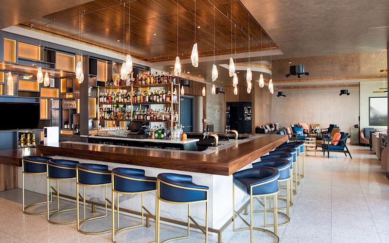 A modern bar with blue stools, hanging lights, and a well-stocked shelf of bottles in a stylish interior seating area.