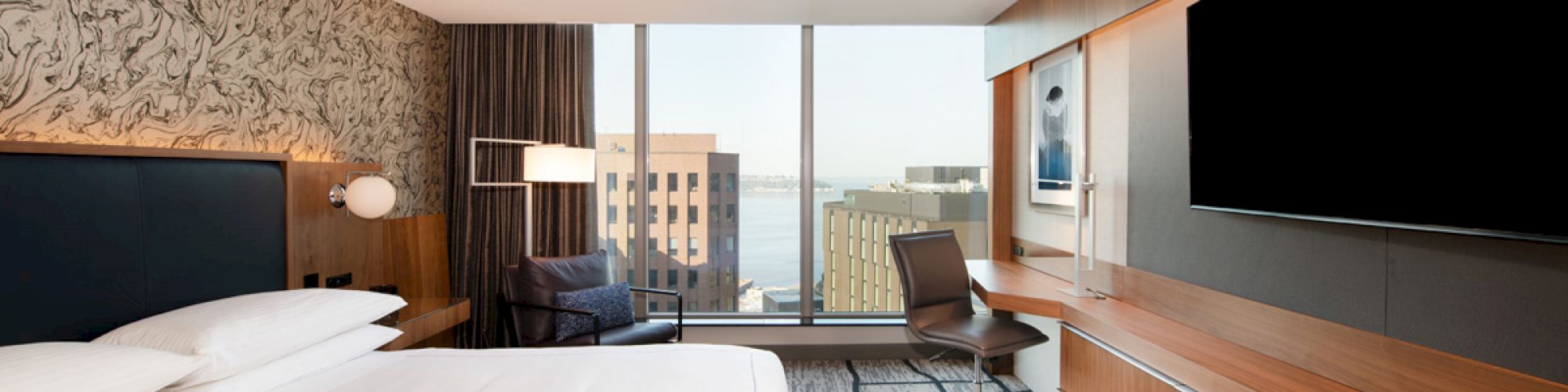 Modern hotel room with a large bed, desk, TV, lounge chair, and window offering a view of city buildings and water.