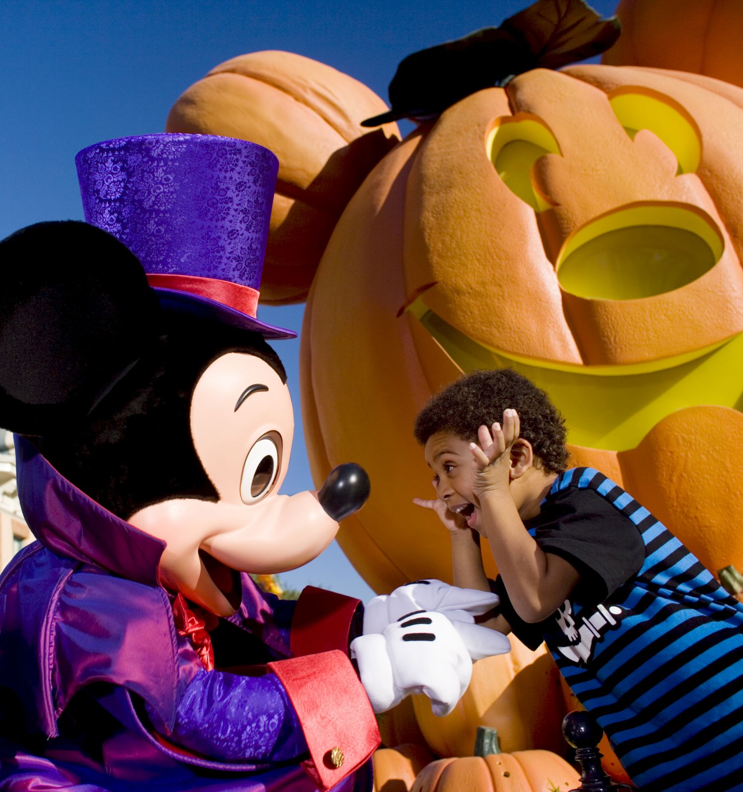 A child excitedly interacts with a character in a Halloween-themed setting, featuring a large, carved pumpkin decoration in the background.