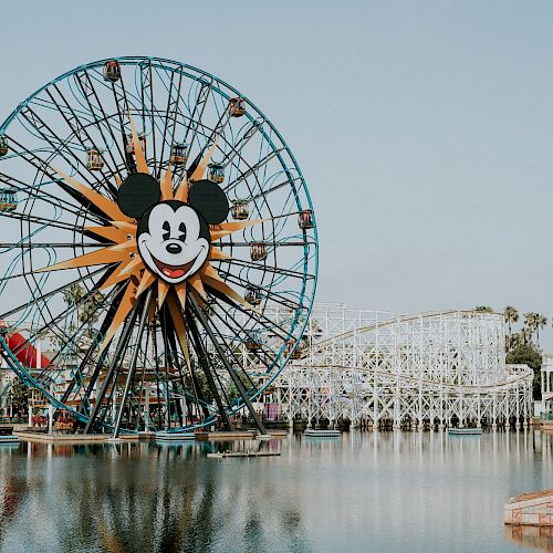 An amusement park with a large Ferris wheel featuring a mouse head, a roller coaster, and various rides are seen across a body of water.