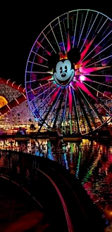 The image shows a large, illuminated Ferris wheel with a design featuring a well-known cartoon character's face against a nighttime amusement park backdrop.